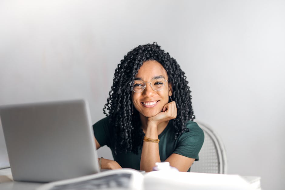 Joyful businesswoman with curly hair smiling at camera while using laptop indoors to study Online Masters' Degree with Highstone Global University.